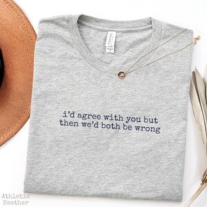 May include: A heather gray crew neck t-shirt featuring the embroidered black text "i'd agree with you but then we'd both be wrong" centered on the chest. The shirt has a white interior neck label and is paired with a delicate gold chain necklace.