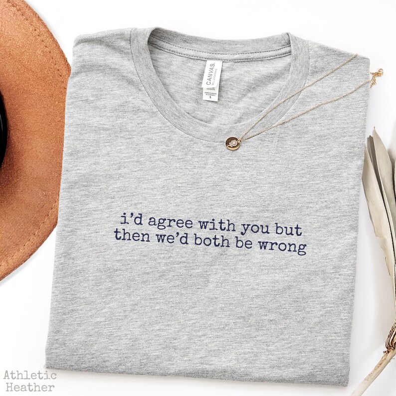 May include: A heather gray crew neck t-shirt featuring the embroidered black text "i'd agree with you but then we'd both be wrong" centered on the chest. The shirt has a white interior neck label and is paired with a delicate gold chain necklace.