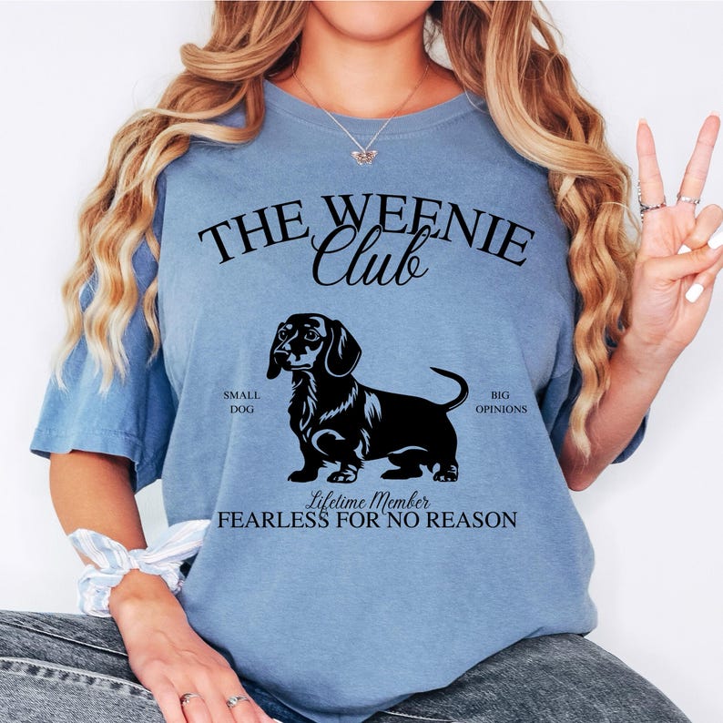May include: Light blue t-shirt with the text "THE WEENIE Club" and a black dachshund graphic. The shirt also includes the phrases "SMALL DOG", "BIG OPINIONS", and "LIFETIME MEMBER FEARLESS FOR NO REASON".