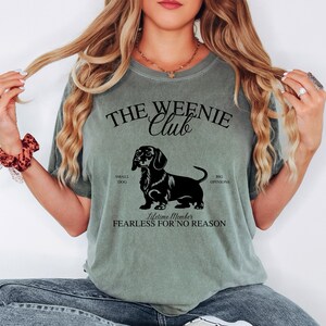May include: Sage green t-shirt with a black graphic of a dachshund and the text "THE WEENIE CLUB". The graphic includes the words "SMALL DOG", "BIG OPINIONS", and "FEARLESS FOR NO REASON".