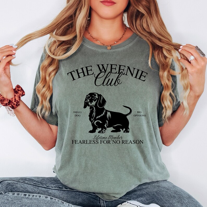 May include: Sage green t-shirt with a black graphic of a dachshund and the text "THE WEENIE CLUB". The graphic includes the words "SMALL DOG", "BIG OPINIONS", and "FEARLESS FOR NO REASON".