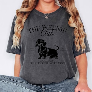 May include: Dark gray t-shirt with the text "THE WEENIE Club" above a black dachshund illustration. The shirt also includes the phrases "SMALL DOG BIG OPINIONS" and "Lifetime Member FEARLESS FOR NO REASON".