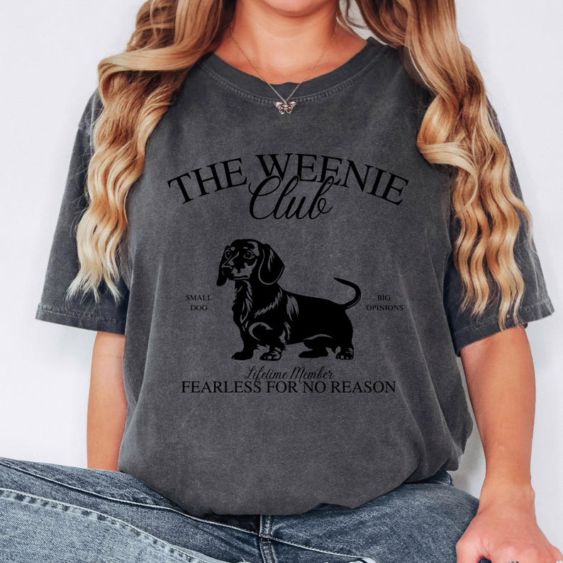 May include: Dark gray t-shirt with the text "THE WEENIE Club" above a black dachshund illustration. The shirt also includes the phrases "SMALL DOG BIG OPINIONS" and "Lifetime Member FEARLESS FOR NO REASON".