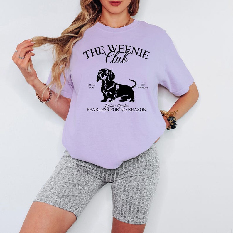 May include: A light purple t-shirt with the text "THE WEENIE Club" above a black dachshund illustration. Below the illustration, the text reads "Lifetime Member FEARLESS FOR NO REASON". The shirt also has the phrases "SMALL DOG" and "BIG OPINIONS".