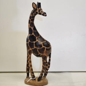 May include: A wooden giraffe sculpture with a long neck and brown and black spotted pattern. The giraffe stands on a small, round, wooden base. The sculpture is hand-carved and has a natural wood finish.