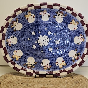 May include: Oval ceramic serving platter with a blue speckled interior and a border of snowmen and snowflakes. The rim has a red and white checkered pattern. A decorative piece for holiday entertaining.