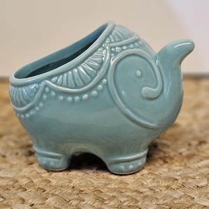 May include: A light blue ceramic elephant planter with a detailed design. The planter has a large opening for plants and is supported by three small feet. The elephant's trunk is curled upwards, and the body is decorated with embossed patterns.