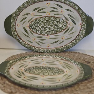 May include: Two oval ceramic baking dishes with handles, featuring a cream-colored base decorated with green leaf patterns, small white floral clusters, and tiny yellow dots. The rims are bordered by a dense green and white patterned band.