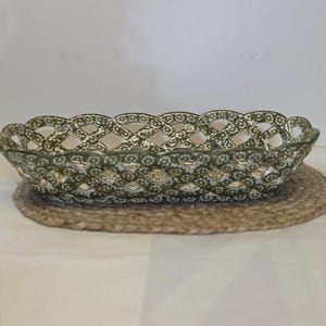 May include: An oval ceramic basket with a lattice-style woven wall design. The exterior features a repeating olive green and white floral pattern across the entire surface, including the scalloped top edge and the open-work sides.