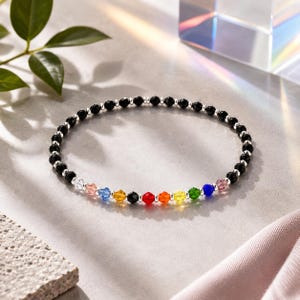 May include: A beaded bracelet featuring a continuous loop of small black faceted beads interspersed with silver-toned spacer beads. A segment of the bracelet displays a sequence of multicolored faceted beads in light pink, blue, yellow, black, red, orange, yellow, green, blue, and pink.