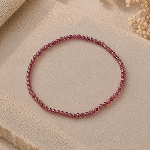 May include: A delicate circular bracelet composed of small, faceted deep red garnet beads. The beads are uniform in size and arranged in a continuous loop, reflecting light across their polished surfaces to create a subtle, shimmering effect.