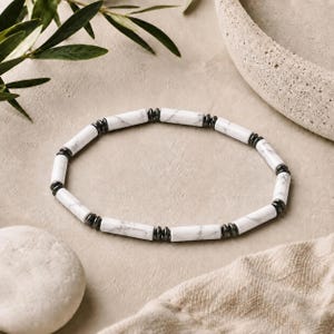 May include: A beaded bracelet featuring alternating white cylindrical beads with grey veining and small, dark metallic disc-shaped spacers. The beads are arranged in a circular pattern, creating a simple, uniform design with a polished finish.