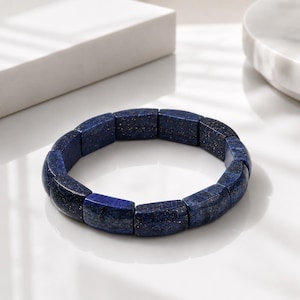 May include: A stretch bracelet composed of rectangular, polished lapis lazuli beads. The beads are a deep, saturated blue color with visible natural gold-colored pyrite flecks distributed throughout the stone surfaces in a repeating pattern.