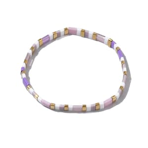 May include: A beaded bracelet featuring a repeating pattern of rectangular beads in white, lavender, light purple, and gold. The bracelet is designed to be worn around the wrist and is set against a white background.