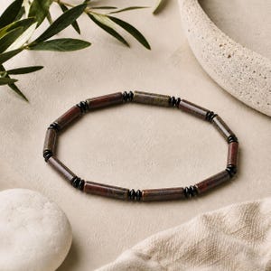 May include: A circular bracelet composed of alternating cylindrical brown stone beads and small, round, glossy black spacer beads. The brown beads feature subtle mottled textures in shades of reddish-brown and dark olive, arranged in a repeating pattern.