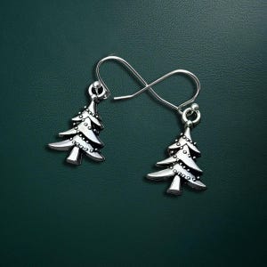 May include: A pair of silver-colored Christmas tree earrings. Each earring features a detailed tree design with small, round accents. The earrings hang from silver-colored ear wires, set against a dark green background.