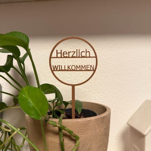 Könnte beinhalten: Ein braunes Holzschild mit dem Text "Herzlich Willkommen" in einem Kreis.
