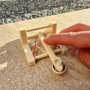 Könnte beinhalten: Eine kleine, beige Holzkatapult mit einem roten Gummiband und einem kleinen Stein in der Schaufel. Die Katapult steht auf einer hellen Oberfläche.