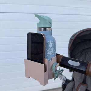 Op de afbeelding: Een roze en lichtblauwe kinderwagenorganizer, met een blauwe waterfles met schelpmotief, een smartphone en een fopspeen. De organizer wordt aan de kinderwagenhandgreep bevestigd.