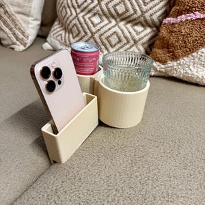 Può includere: Un porta telefono e bevande in plastica bianca con un telefono cellulare, una lattina di acqua frizzante e una tazza di vetro. Il supporto è seduto su un divano.