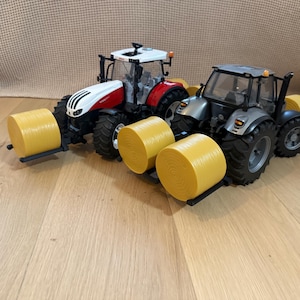 Könnte beinhalten: Zwei Spielzeugtraktoren mit Heuballen, die hinten befestigt sind. Der Traktor links ist weiß und rot, während der Traktor rechts grau ist. Beide Traktoren haben schwarze Räder.