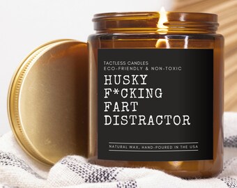 May include: A lit, amber-colored glass jar candle with a gold lid. The black label reads "HUSKY F*CKING FART DISTRACTOR" in white text. The candle is labeled "TACTLESS CANDLES ECO-FRIENDLY & NON-TOXIC" and "NATURAL WAX, HAND-POURED IN THE USA."