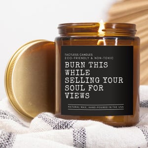 May include: A brown glass candle with a black label that reads "TACTLESS CANDLES ECO-FRIENDLY & NON-TOXIC BURN THIS WHILE SELLING YOUR SOUL FOR VIEWS NATURAL WAX, HAND-POURED IN THE USA".