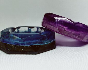 以下が含まれることがあります： 幾何学的な樹脂製の灰皿が2つ。1つは黒のアクセントがついた濃い青で、もう1つは明るい紫です。
