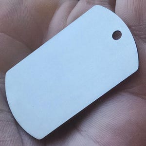 Könnte beinhalten: Ein leerer, rechteckiger Erkennungsmarke mit abgerundeten Ecken und einem einzelnen Loch oben. Die Marke hat eine helle, neutrale Farbe, möglicherweise Silber oder Weiß, und scheint aus Metall zu sein. Die Marke wird in einer Hand gehalten.