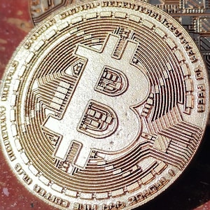 May include: Three gold Bitcoin coins are shown, each with detailed designs. The coins display the Bitcoin symbol and circuit-board patterns. They are arranged on a reddish-brown surface, with shadows adding depth to the image.