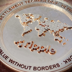 May include: Close-up of a silver coin with the word "Ripple" embossed in the center. The coin features a world map and the text "Value Without Borders" and "Settlement Technology Ultra Work" around the edge.