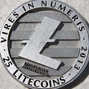 May include: A silver Litecoin coin with the Litecoin symbol in the center. The coin is marked with "25" and "2013" and the text "VIRES IN NUMERIS" and "LITECOINS". The coin is marked "TROY OUNCE .999 FINE SILVER".