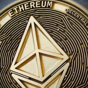 May include: Close-up of a gold Ethereum coin. The coin features the word "ETHEREUM" at the top and a geometric symbol in the center. The coin has a detailed, textured design with the word "DECENTRAL" visible.