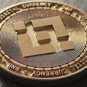 Peut inclure: Gros plan d'une pièce de monnaie numérique Binance Coin (BNB) dorée. La pièce présente le logo Binance et les mots "DIGITAL CURRENCY" et "BNB" sur les bords. La pièce a un design texturé et circulaire.