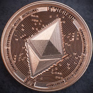Könnte beinhalten: Nahaufnahme einer kupferfarbenen Ethereum-Münze. Die Münze zeigt das Ethereum-Logo, eine stilisierte Diamantenform, in der Mitte. Der Rand der Münze trägt die Worte "DECENTRALIZED APPLICATION SMART CONTRACTS" und "ETHEREUM".