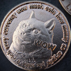 May include: A bronze-colored coin featuring a detailed relief of a dog's face with the word "WOW" prominently displayed. The coin is inscribed with phrases like "very currency" and "much coin," and "plz mine." The coin has a shiny finish.
