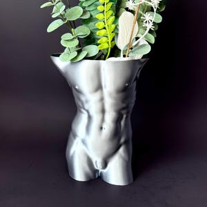 May include: A silver-colored decorative vase shaped like a male torso. The vase has detailed musculature and holds a variety of green and white artificial plants. The vase is set against a dark background.