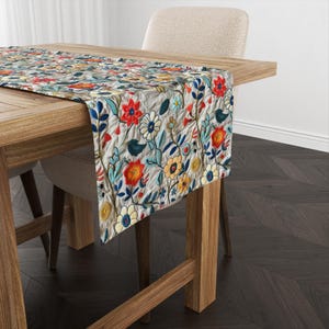 May include: A floral table runner with a white background and colorful flowers in shades of red, blue, yellow, and green. The runner is placed on a wooden table with a chair.