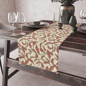 May include: A cream-coloured table runner featuring a repeating pattern of red leaves and green vines. The runner is laid on a dark wooden table, set with plates, glasses, and decorative items.
