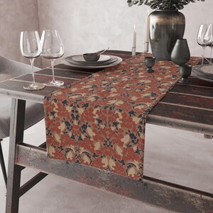 Könnte beinhalten: Ein Tischläufer mit floralem Muster in Rot, Beige und Marineblau. Er liegt auf einem dunklen Holztisch, mit Weingläsern und Geschirr. Das Design zeigt ein sich wiederholendes Muster aus Blumen und Blättern.