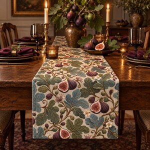 May include: A rectangular table runner featuring a woven pattern of green fig leaves, purple figs, and small white flowers on a light beige background. The fabric displays a detailed botanical design with intricate textures and natural color tones.