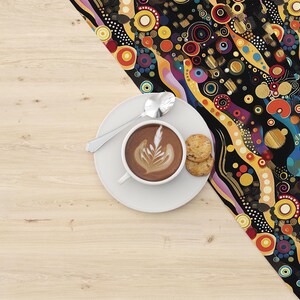 Puede incluir: Una taza de cerámica blanca con un diseño de arte latte, un plato con dos galletas y una cuchara sobre una mesa de madera con una tela estampada colorida en el fondo.