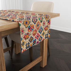 May include: A colorful table runner with a geometric pattern of embroidered flowers and squares on a beige background. The runner is on a wooden table with a chair.