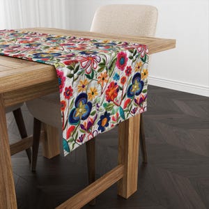 Könnte beinhalten: Ein weißer Tischläufer mit einem bunten floralen Stickmuster. Der Läufer zeigt eine Vielzahl von Blumen in Rot-, Rosa-, Gelb-, Blau- und Grüntönen. Der Läufer liegt auf einem Holztisch mit einem dunklen Holzboden.
