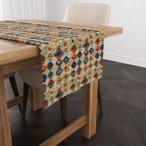 May include: A beige table runner with a colorful geometric pattern. The pattern features a diamond shape with embroidered details in shades of red, orange, yellow, green, blue, and purple.