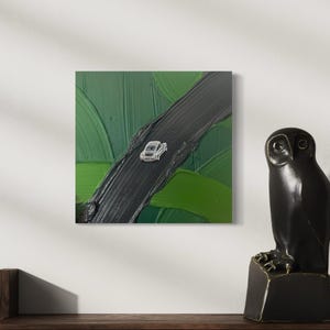 Könnte beinhalten: Ein quadratisches abstraktes Gemälde zeigt ein weißes Auto auf einer schwarzen Straße, vor dem Hintergrund grüner Pinselstriche. Das Kunstwerk ist an einer weißen Wand befestigt. Eine schwarze Eulenstatue steht auf einer Holzoberfläche.