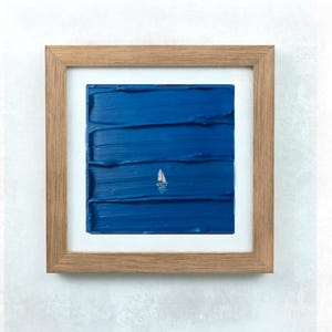 May include: Framed artwork depicting a textured blue sea with a small white sailboat. The painting is set within a light brown wooden frame and a white mount.