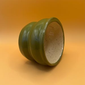 Könnte beinhalten: Ein grüner Keramiktopf mit einem gesprenkelten beigefarbenen Innenraum. Der Topf hat eine abgerundete Form und eine glatte, glänzende Oberfläche.