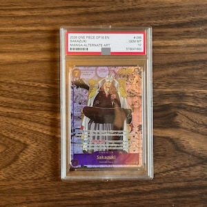May include: A graded One Piece trading card featuring Sakazuki in a manga alternate art style. The card is encased in a clear plastic slab with a label at the top reading 2026 ONE PIECE OP16 EN SAKAZUKI MANGA ALTERNATE ART #065 GEM MT 10.