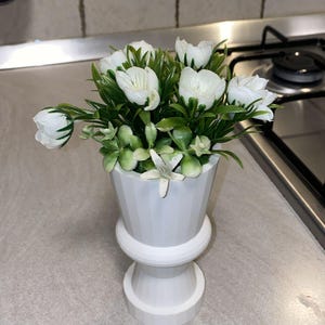 May include: A white, decorative flower pot with a pedestal base, filled with white faux tulips and green foliage. The pot has a geometric design and is placed on a light-colored countertop. The arrangement adds a touch of elegance.